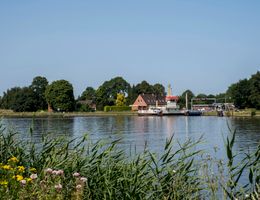 Fähre Breiholz am Ufer eines Flusses mit Häusern und Bäumen im Hintergrund.